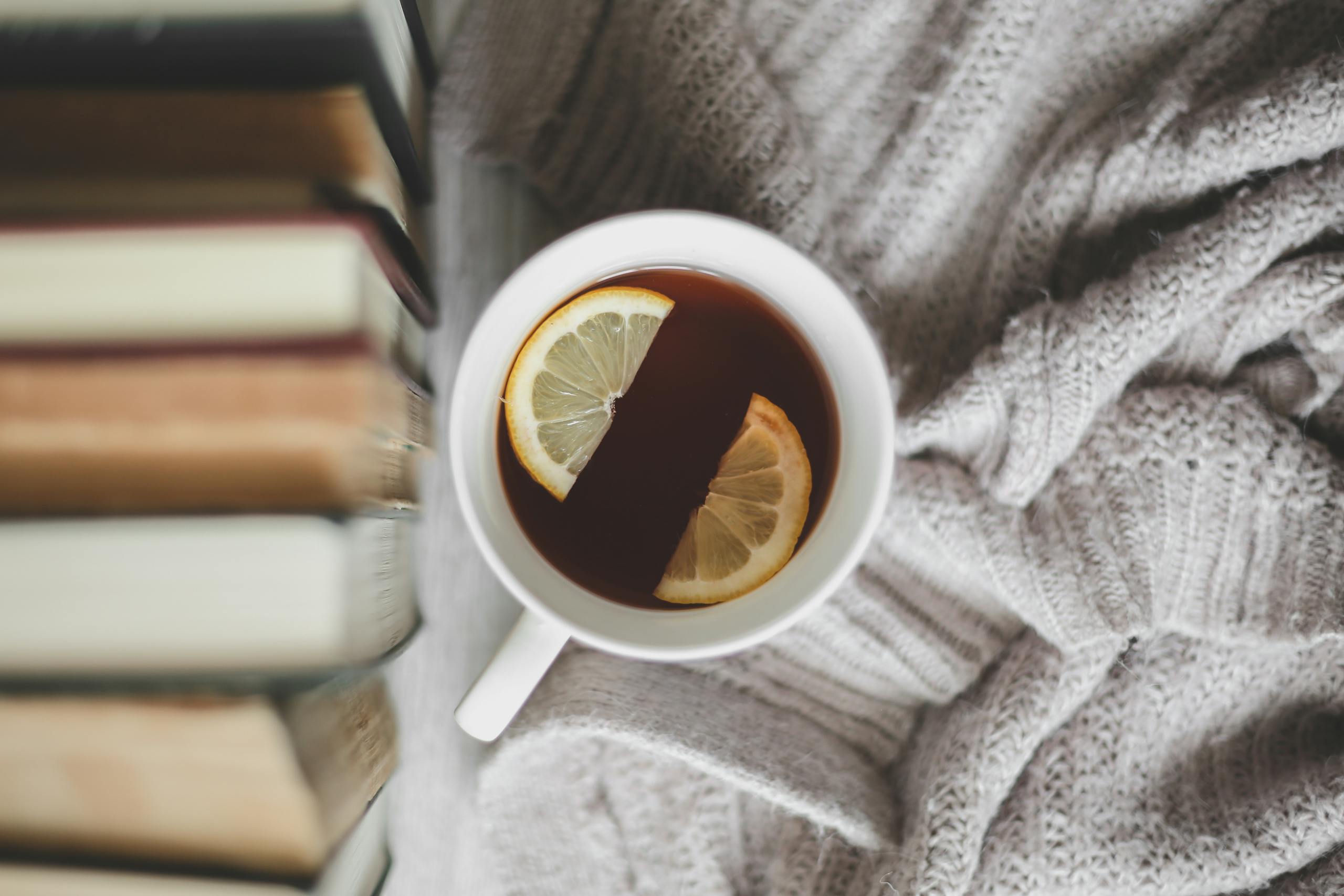 A cup of tea with lemon slices, surrounded by books and a warm sweater for a cozy indoors vibe.